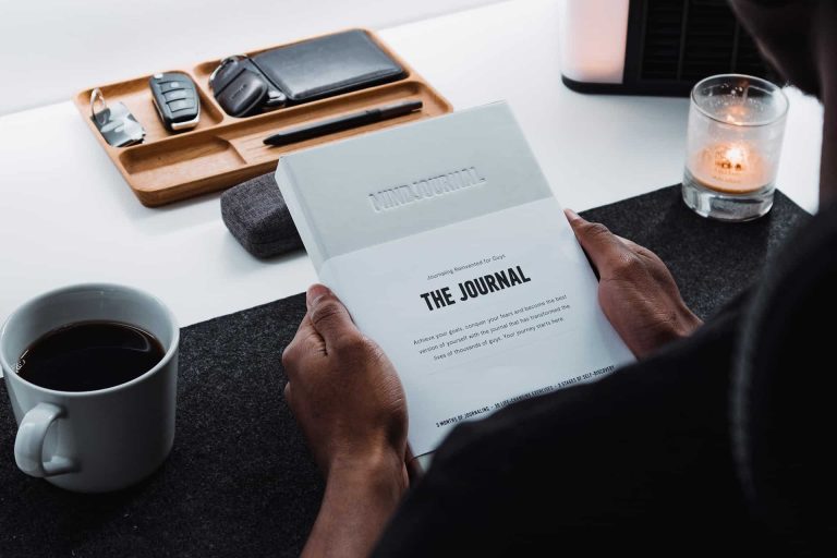 A person holds a white journal at a desk with a cup of coffee, a lit candle, and a tray holding keys and a wallet—planning goals or tracking foundation funding opportunities.