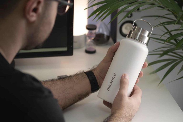 A person holding a white refillable water bottle labeled alliance sits at a desk, surrounded by a plant and computer monitor, reflecting on Foundation Funding opportunities.