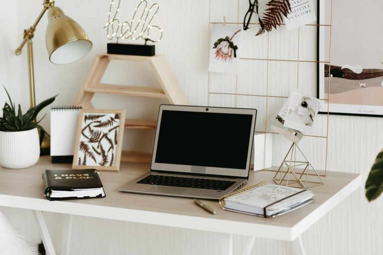 A tidy desk with a laptop, notebooks, a pen, greeting card about grants funding, lamp, small plant, and decorative items, set against a white background with wire grid and framed pictures.