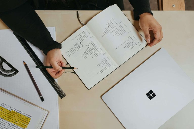 Person writing in a notebook filled with graphs and notes, next to a closed laptop, ruler, protractor, pencil, and a sheet with highlighted text—perfect for organizing Foundation Funding or Grants Funding proposals.