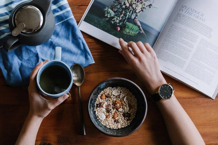 A person reads a magazine while holding a mug of tea, with a bowl of cereal, a spoon, and a teapot on a wooden table—perhaps exploring Foundation Funding or Grants Funding opportunities inside.
