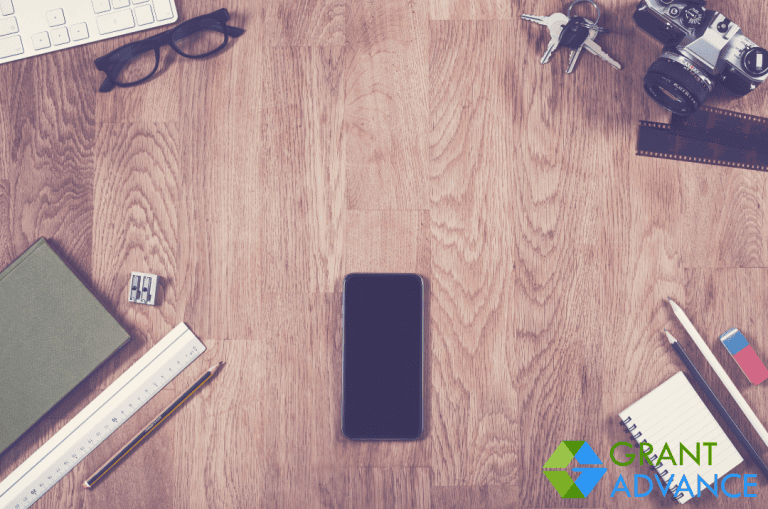 A smartphone sits on a wooden desk surrounded by glasses, camera, keys, stationery, a notepad, and the Grant Advance logo in the bottom right corner—perfect for exploring top Grant Search Engines.