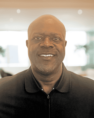 A man with a shaved head and a short beard smiles at the camera in a dark zip-up collared shirt, set against a blurred indoor background—ready to discuss foundation funding and grants funding opportunities.