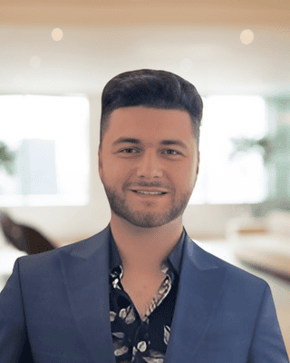 A man with short blond hair and a trimmed beard, wearing a dark blazer and black shirt, smiles at the camera in a bright, modern indoor setting, reflecting confidence often seen among professionals in Grants Funding.
