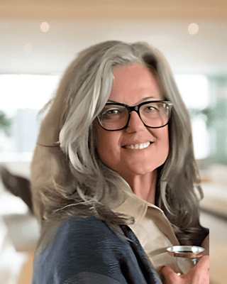 A woman with gray hair and glasses smiles while holding a glass indoors in a modern, brightly lit room, celebrating recent grants funding success.