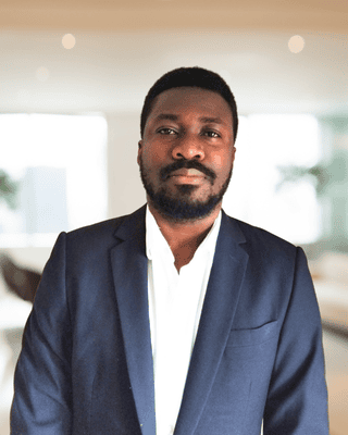 Meet our Experts: A man in a dark suit jacket and white shirt stands indoors in a well-lit, modern room with a blurred background.